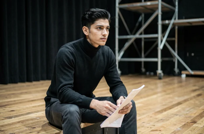 Joven actor sentado en un escenario de teatro negro, sosteniendo un guion, mirada pensativa en una atmósfera de repetición teatral